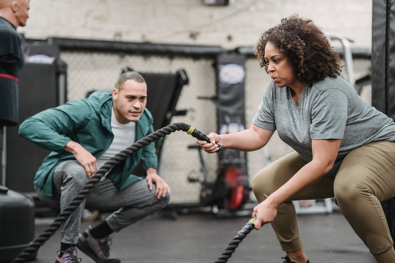 Woman engaged in an intense battle rope workout guided by a coach in a gym setting.